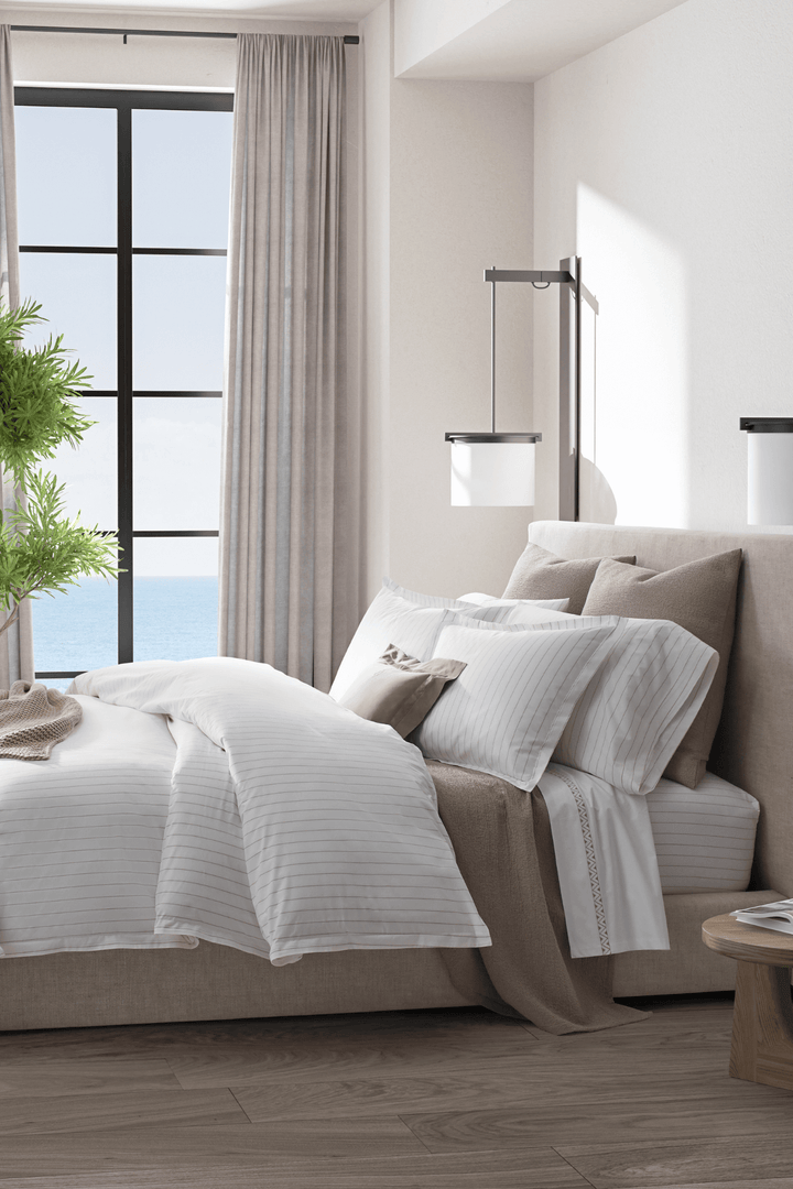 Bedroom with a bed covered in Matouk Amalfi white and brown bedding, and a large window in the background.